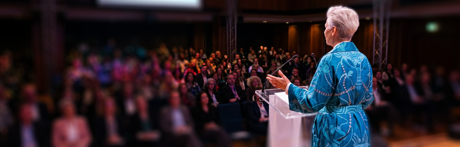 Eine Rednerin steht, von hinten gesehen, am Pult vor einem Publikum, dessen Gesichter unscharf bleiben (KI-generiertes Bild)